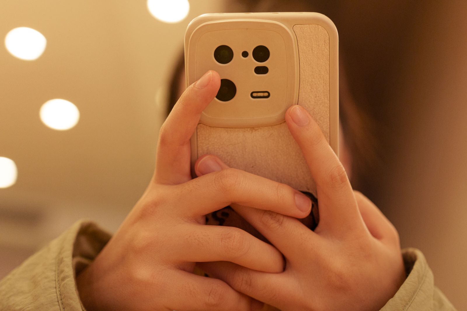 暖かい色の明かりがぼける室内で、ベージュのケースのスマートフォンを両手で縦に持つ手元。こちらに見えているのは背面のカメラ部分