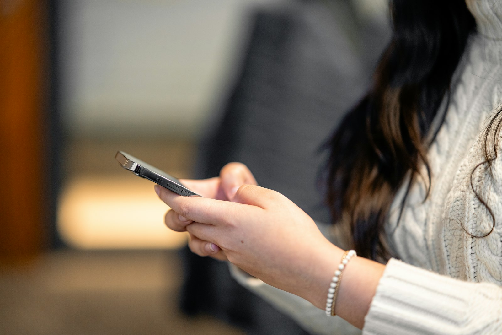 白いケーブル編みのセーターを着た人が、スマートフォンを両手で持って操作している手元を横から写した写真。画面の中身は見えない