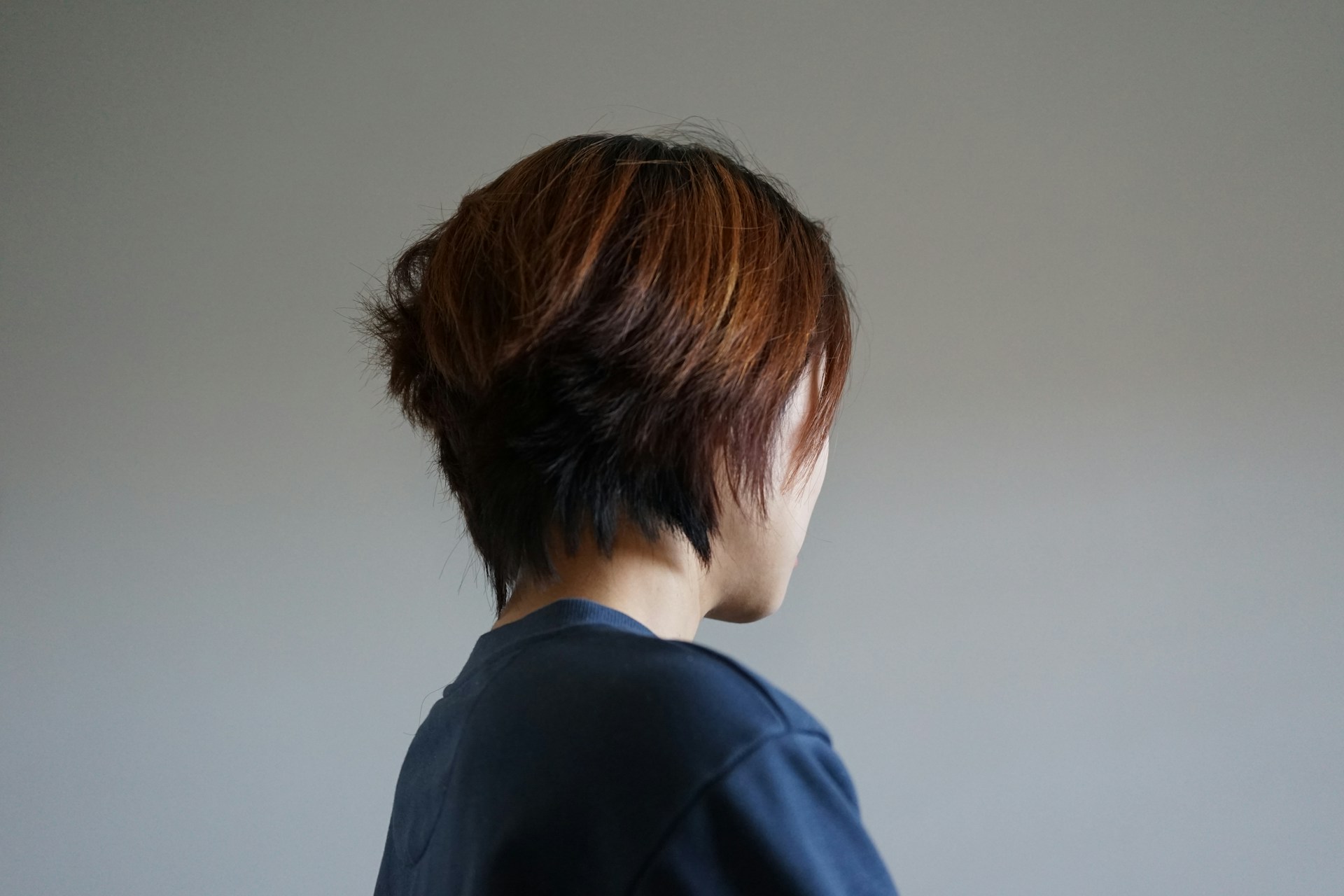 こげ茶の髪に明るい茶色の筋が入った短い髪の人を、斜め後ろから写した写真。紺のシャツを着ていて、背景は灰色の無地。顔は写っていない