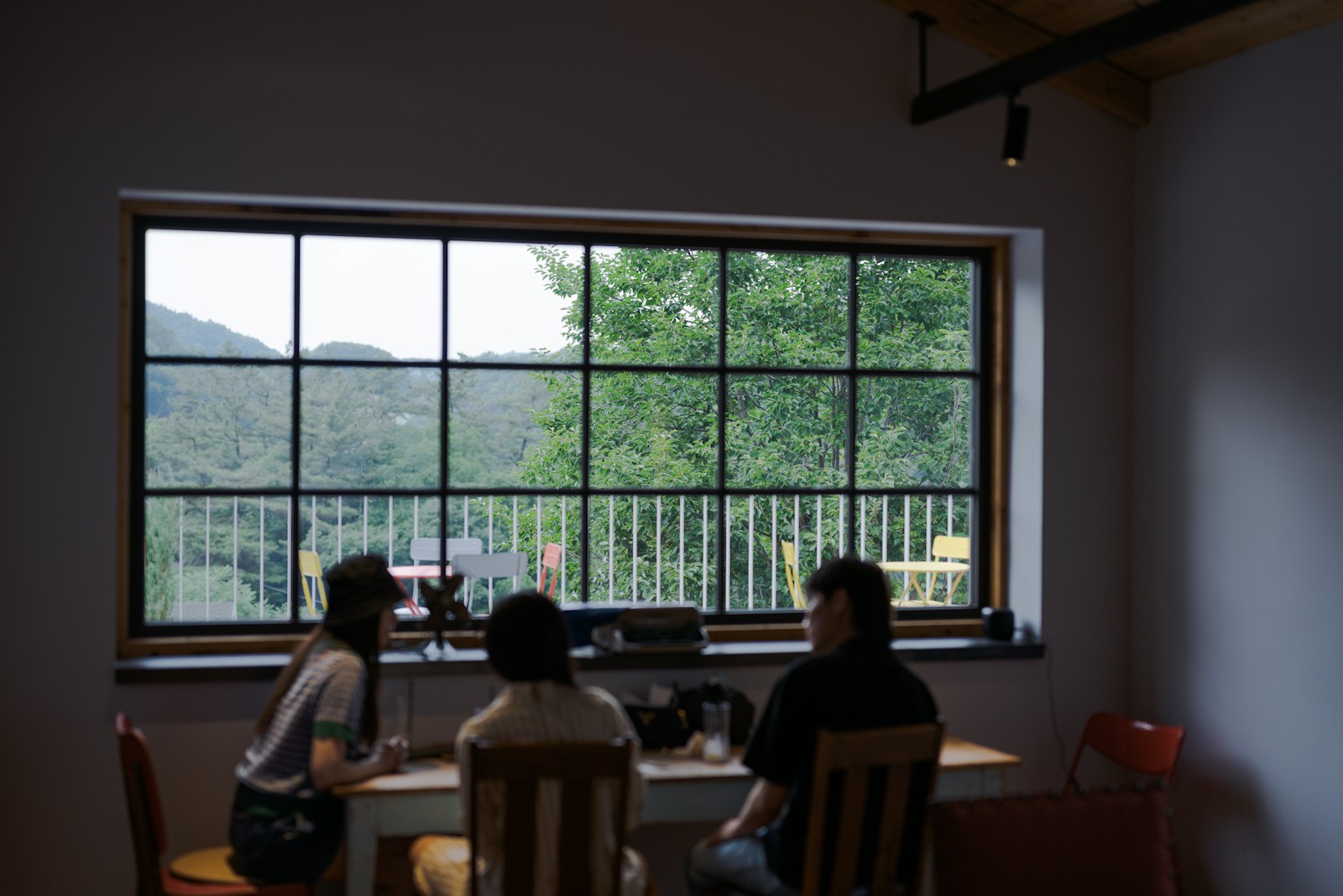 大きな格子窓の前の木のテーブルに3人が座っているカフェの店内。人はぼけていて顔は分からない。窓の外に木々と山、テラスに黄色と赤の椅子が見える