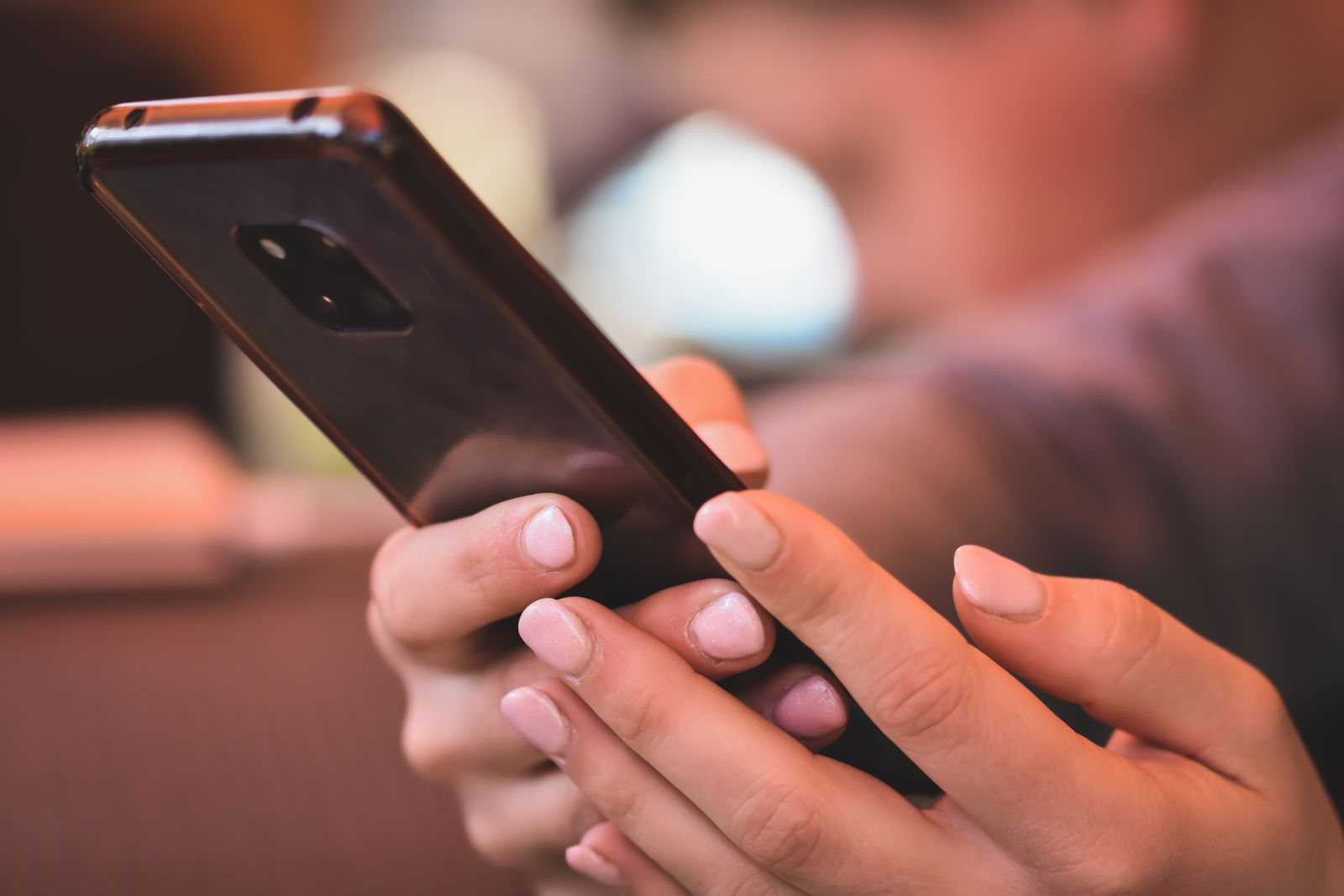 両手でスマートフォンを持っている手元。スマートフォンの背面が見え、背景は暖かい色の室内で大きくぼけている