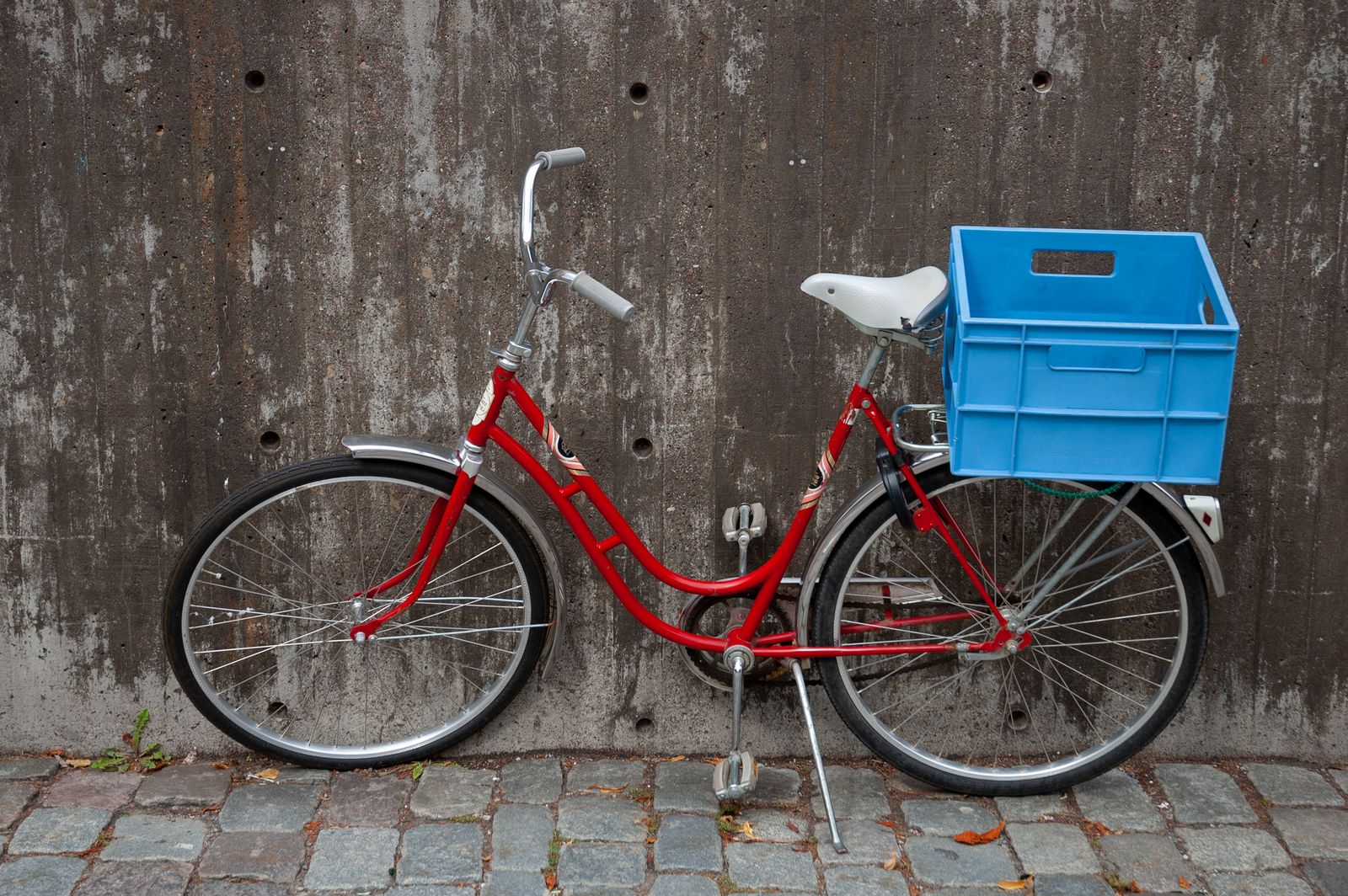 灰色のコンクリートの壁に立てかけた赤い自転車。荷台に青いプラスチックの箱が載っている